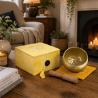 A yellow singing bowl set with a wooden mallet and matching box, styled on a wooden table in a cozy living room with a fireplace in the background.