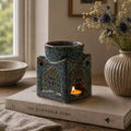 Square Buddha Burner - Dusty Blue ceramic oil burner with Buddha face design, gently warming with a lit tea candle, placed on a book on a wooden surface near dried flowers.