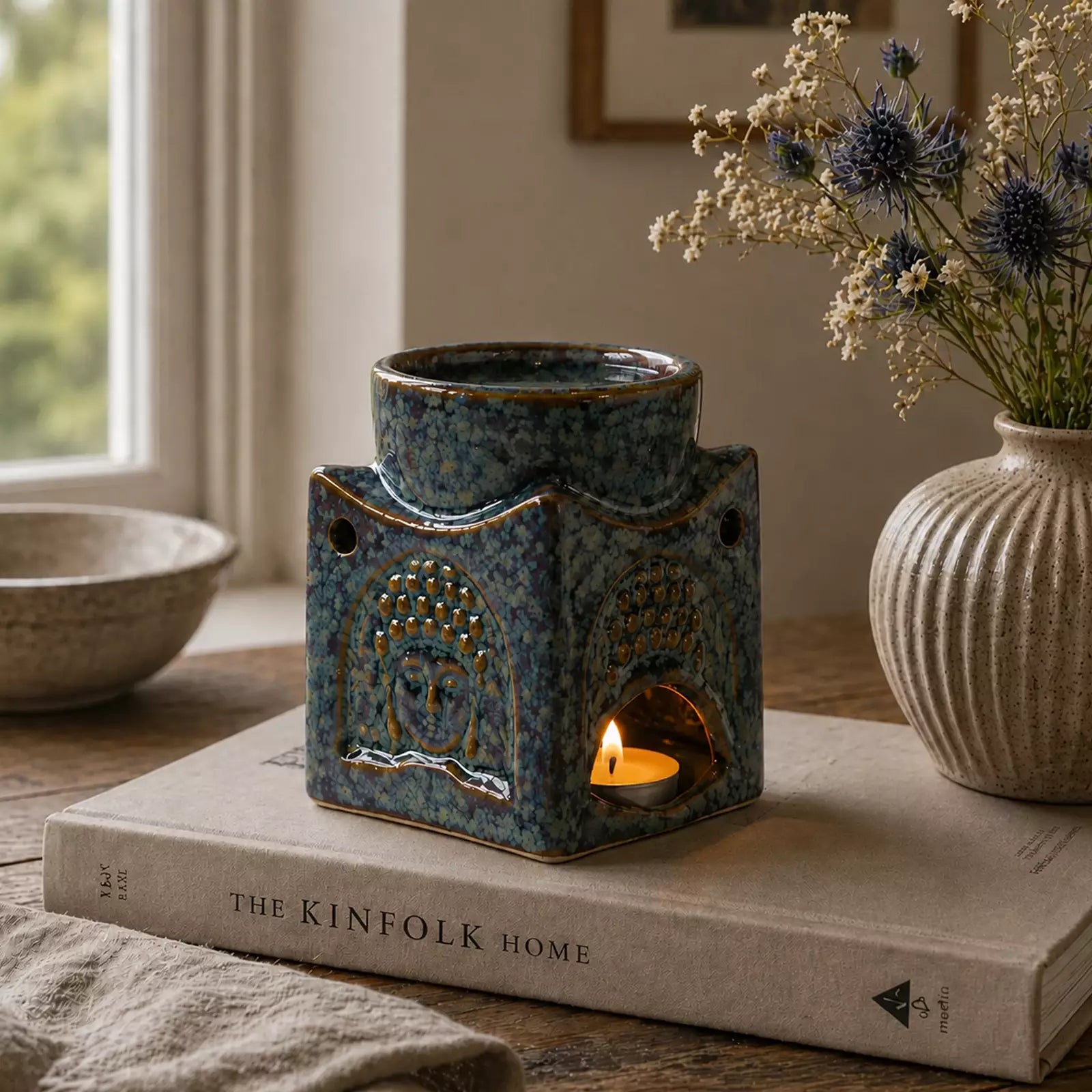 Square Buddha Burner - Dusty Blue ceramic oil burner with Buddha face design, gently warming with a lit tea candle, placed on a book on a wooden surface near dried flowers.