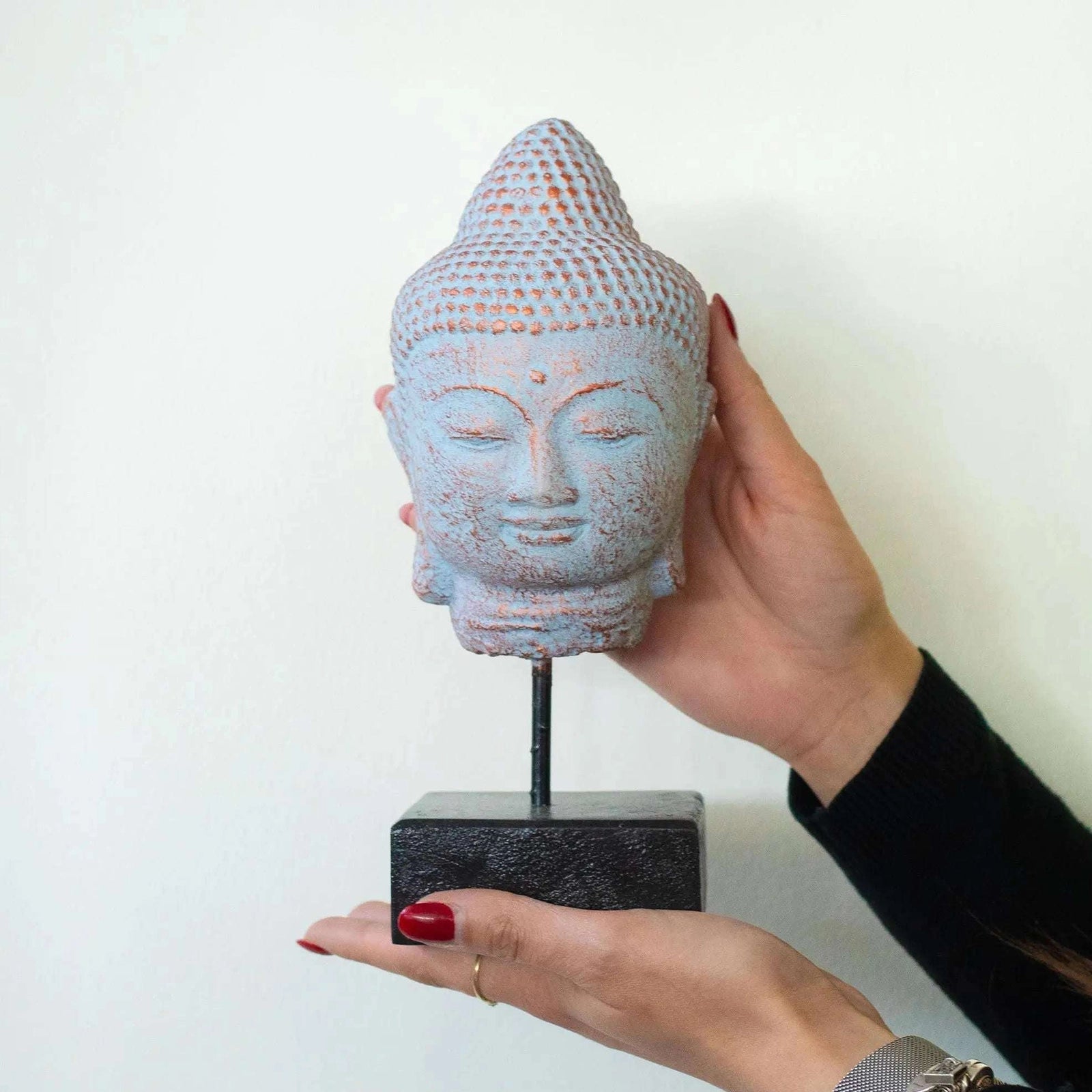 A hand-painted Buddha head sculpture in a light blue-gray color with distressed orange-brown accents, mounted on a black stand and held by two hands against a plain white wall.
