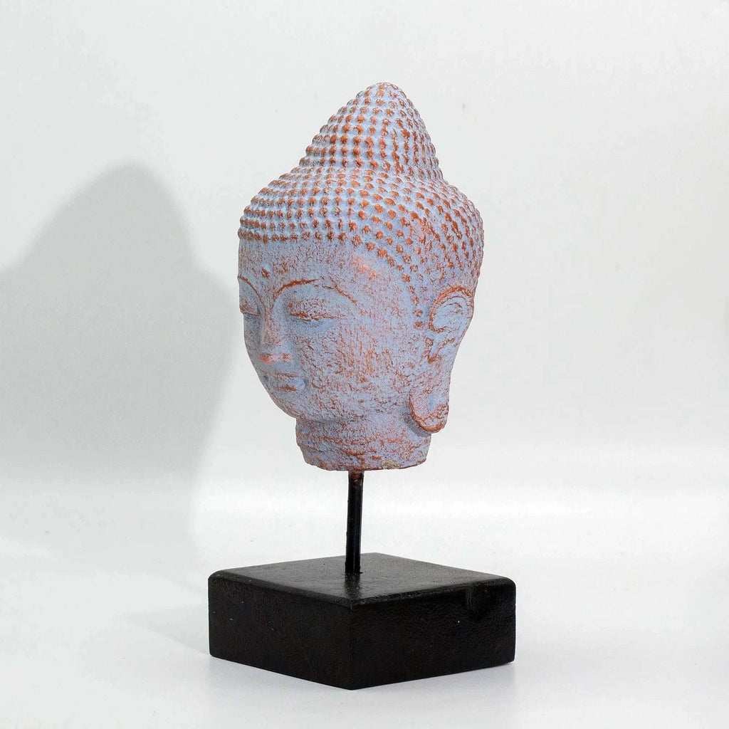 A small Buddha head sculpture with a textured, dotted pattern in pale pinkish-lavender and warm beige tones, mounted on a black metal stand with a square black base against a plain white background.