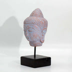 A small Buddha head sculpture with a textured, dotted pattern in pale pinkish-lavender and warm beige tones, mounted on a black metal stand with a square black base against a plain white background.