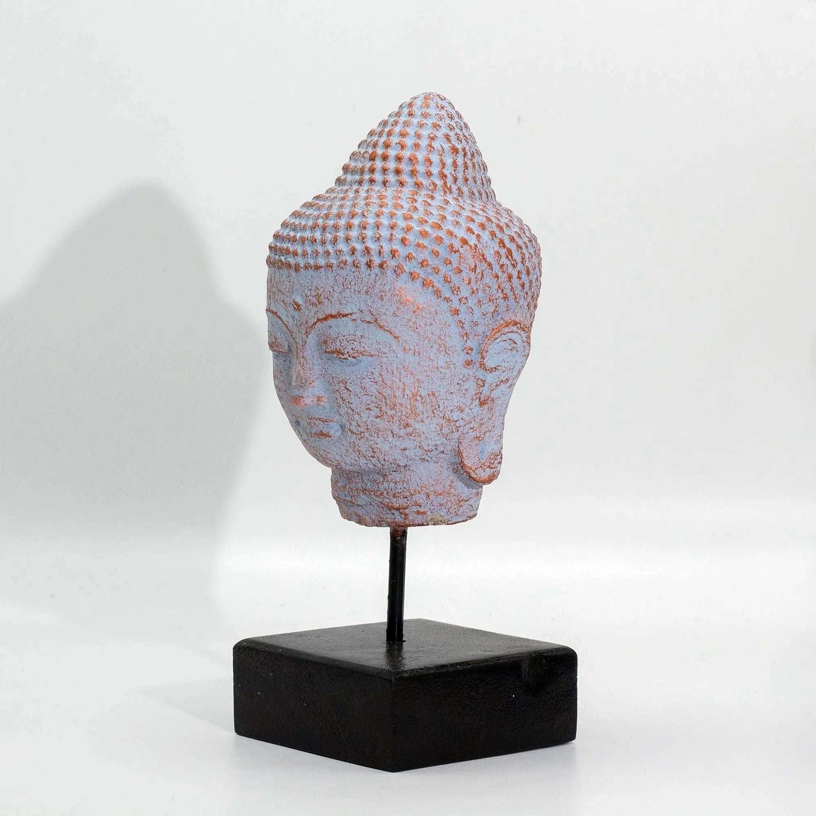 A small Buddha head sculpture with a textured, dotted pattern in pale pinkish-lavender and warm beige tones, mounted on a black metal stand with a square black base against a plain white background.