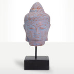 A decorative Buddha head sculpture with a distressed blue and terracotta finish, mounted on a black metal stand with a square black base against a white studio background.