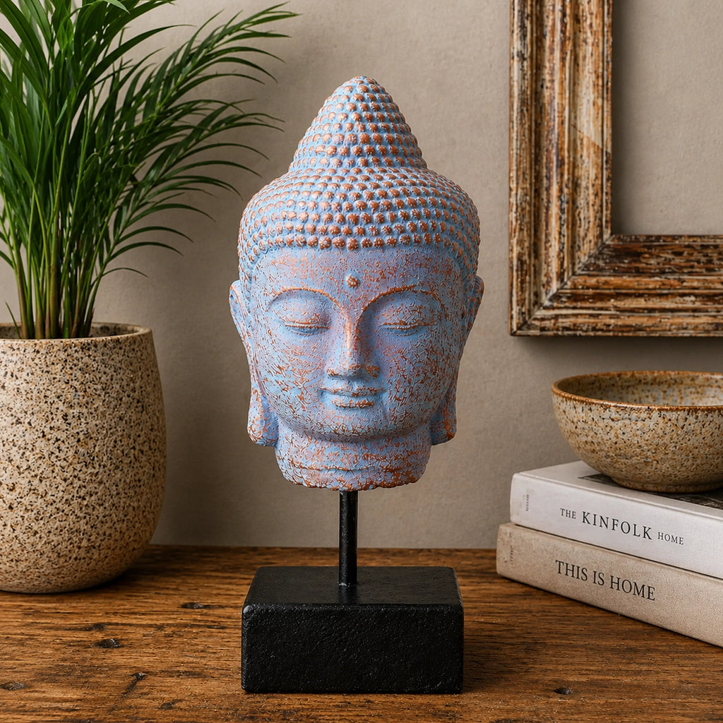 A blue and beige textured Buddha head statue on a black metal stand with a square base, displayed on a wooden table with a potted plant, books, and a bowl in the background.