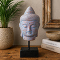 A blue and beige textured Buddha head statue on a black metal stand with a square base, displayed on a wooden table with a potted plant, books, and a bowl in the background.