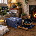 A blue textured gift box with a small cutout on top and a hanging tassel tag sits on a wooden table in a cozy living room. Next to it is a dark blue singing bowl with a gold rim, a wooden mallet, and candles, with a fireplace and armchair in the background.