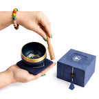 A navy blue singing bowl with a gold rim, held by two hands on a matching navy cushion, with a wooden mallet being struck. A blue presentation box with a tassel and a white geometric logo sits to the right.