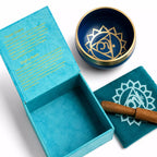 A blue and gold chakra singing bowl set with its turquoise presentation box, a teal fabric cushion printed with a white chakra symbol, and a wooden mallet on a white background.