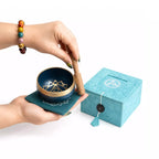 A teal and gold singing bowl set with a wooden mallet and matching teal gift box, shown on a white background with a person's hands holding the bowl.