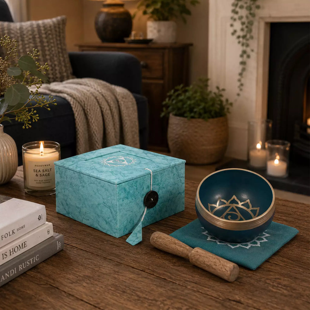 A teal and gold singing bowl set displayed on a wooden table in a cozy living room setting, next to a matching decorative box, a wooden mallet, and lit candles.