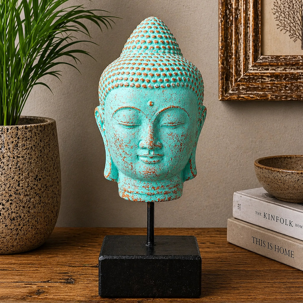 A turquoise Buddha head sculpture with distressed brown patina details, mounted on a black metal stand with a square black base, displayed on a wooden table next to a potted plant and books.