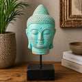 A turquoise Buddha head sculpture with distressed brown patina details, mounted on a black metal stand with a square black base, displayed on a wooden table next to a potted plant and books.