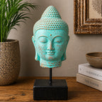A turquoise Buddha head sculpture with distressed brown patina details, mounted on a black metal stand with a square black base, displayed on a wooden table next to a potted plant and books.
