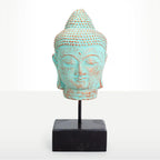A decorative Buddha head sculpture with a distressed turquoise and brown patina finish, mounted on a black metal stand with a square black base, set against a plain white background.