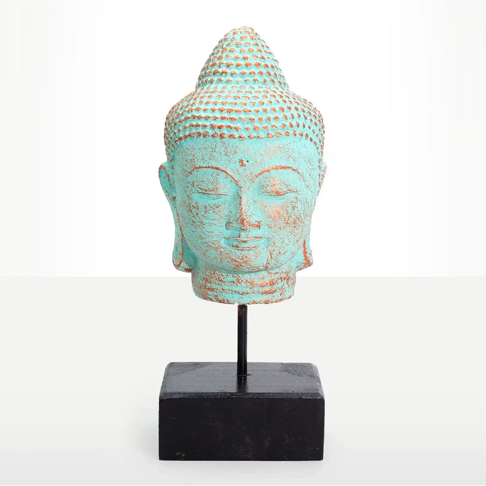 A decorative Buddha head sculpture with a distressed turquoise and brown patina finish, mounted on a black metal stand with a square black base, set against a plain white background.