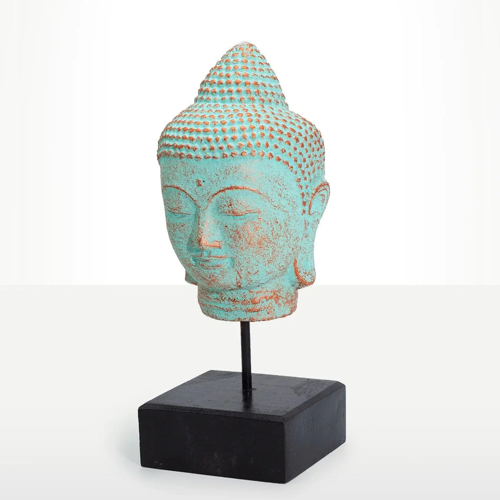 A decorative Buddha head sculpture with a distressed turquoise and rust-colored patina, mounted on a black metal stand with a square black base, set against a clean white studio background.