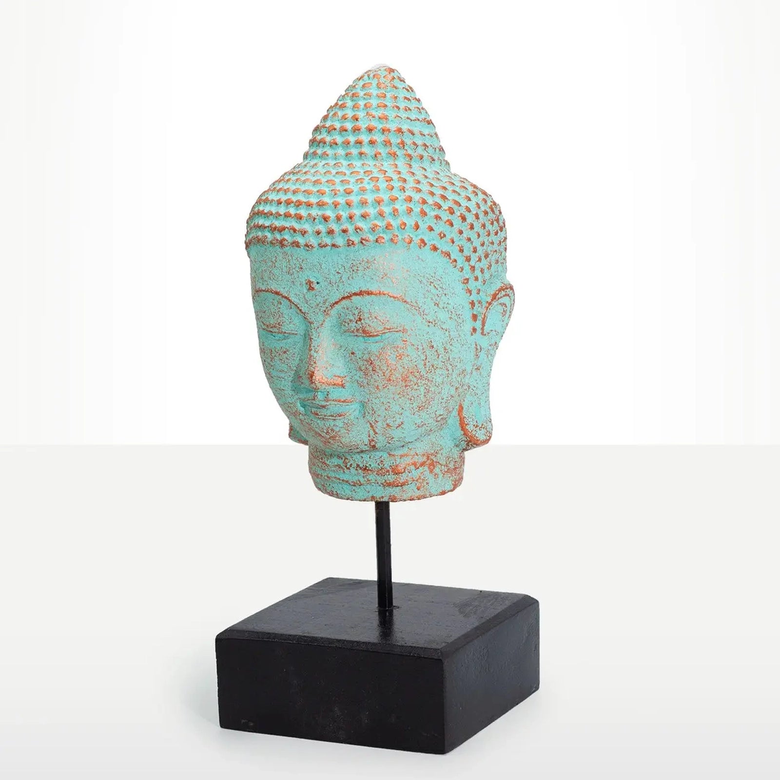 A decorative Buddha head sculpture with a distressed turquoise and rust-colored patina, mounted on a black metal stand with a square black base, set against a clean white studio background.