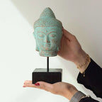 A hand-painted turquoise and brown distressed Buddha head sculpture on a black stand, held by two hands against a plain white background.