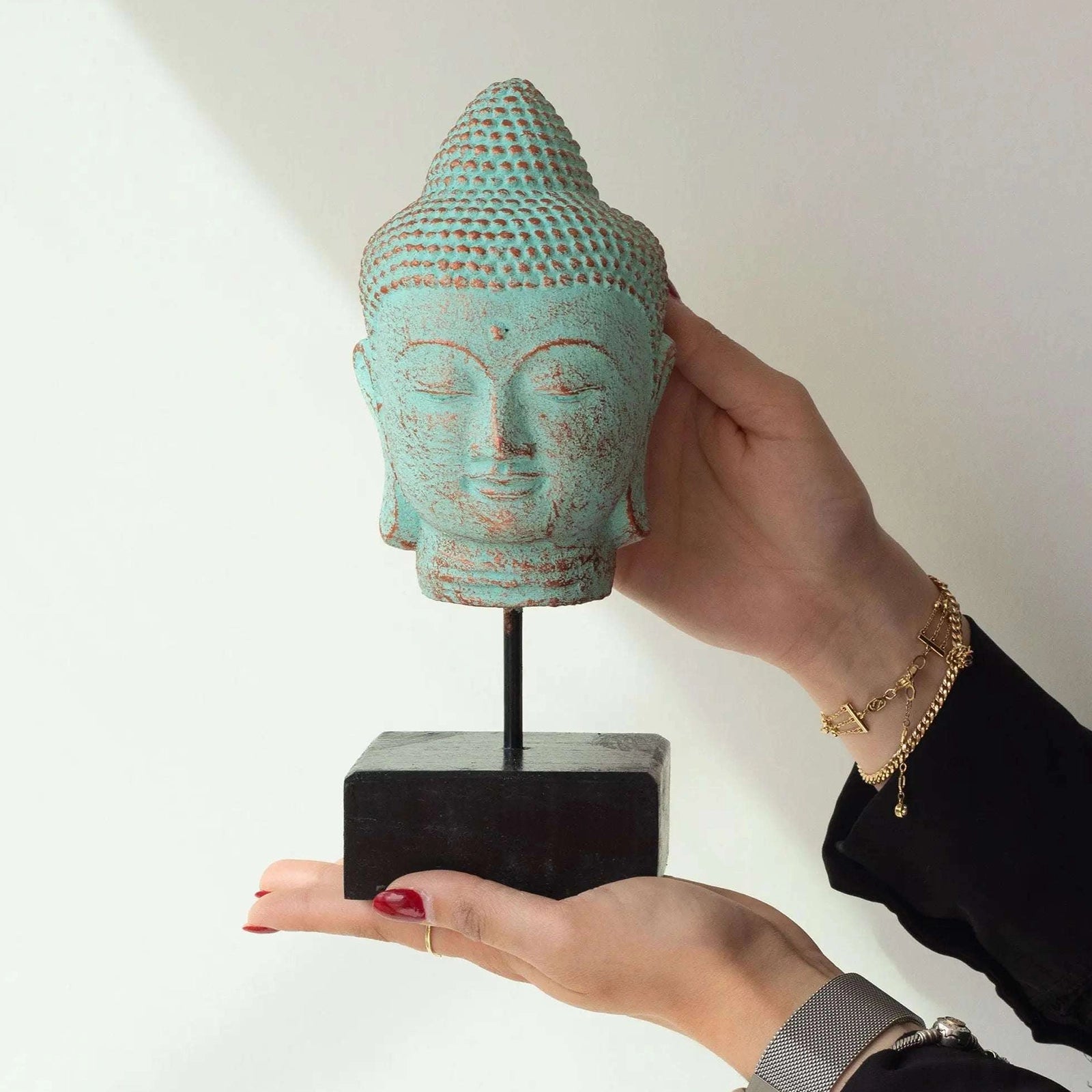 A hand-painted turquoise and brown distressed Buddha head sculpture on a black stand, held by two hands against a plain white background.