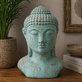 A distressed turquoise blue Buddha bust sculpture with a weathered brown patina, displayed on a wooden shelf next to a potted plant and decorative books.