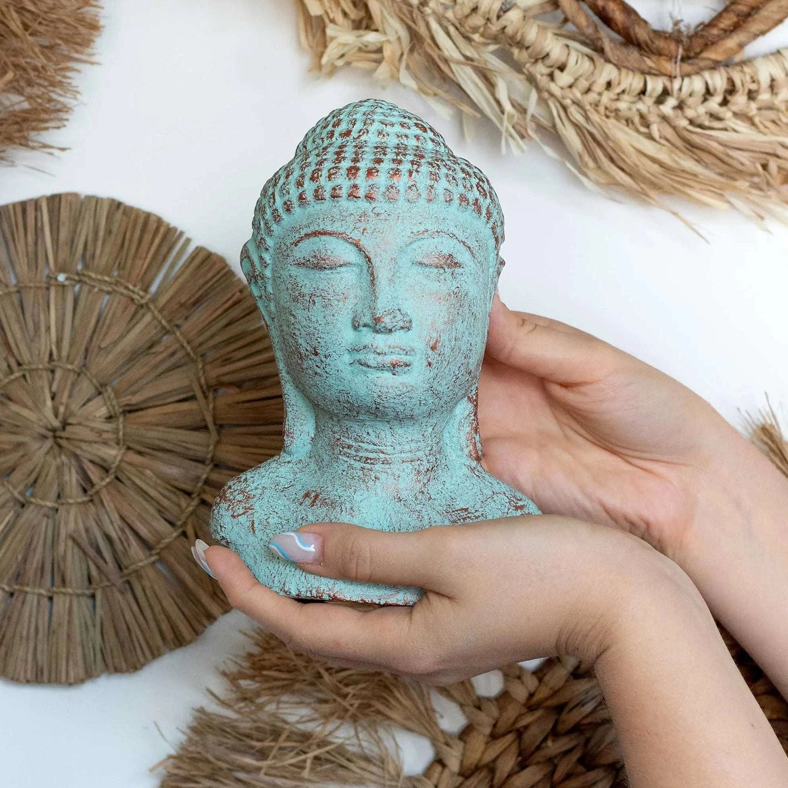 A small Buddha head bust sculpture with a distressed turquoise and brown patina finish, held in two hands against a white background decorated with woven straw elements.