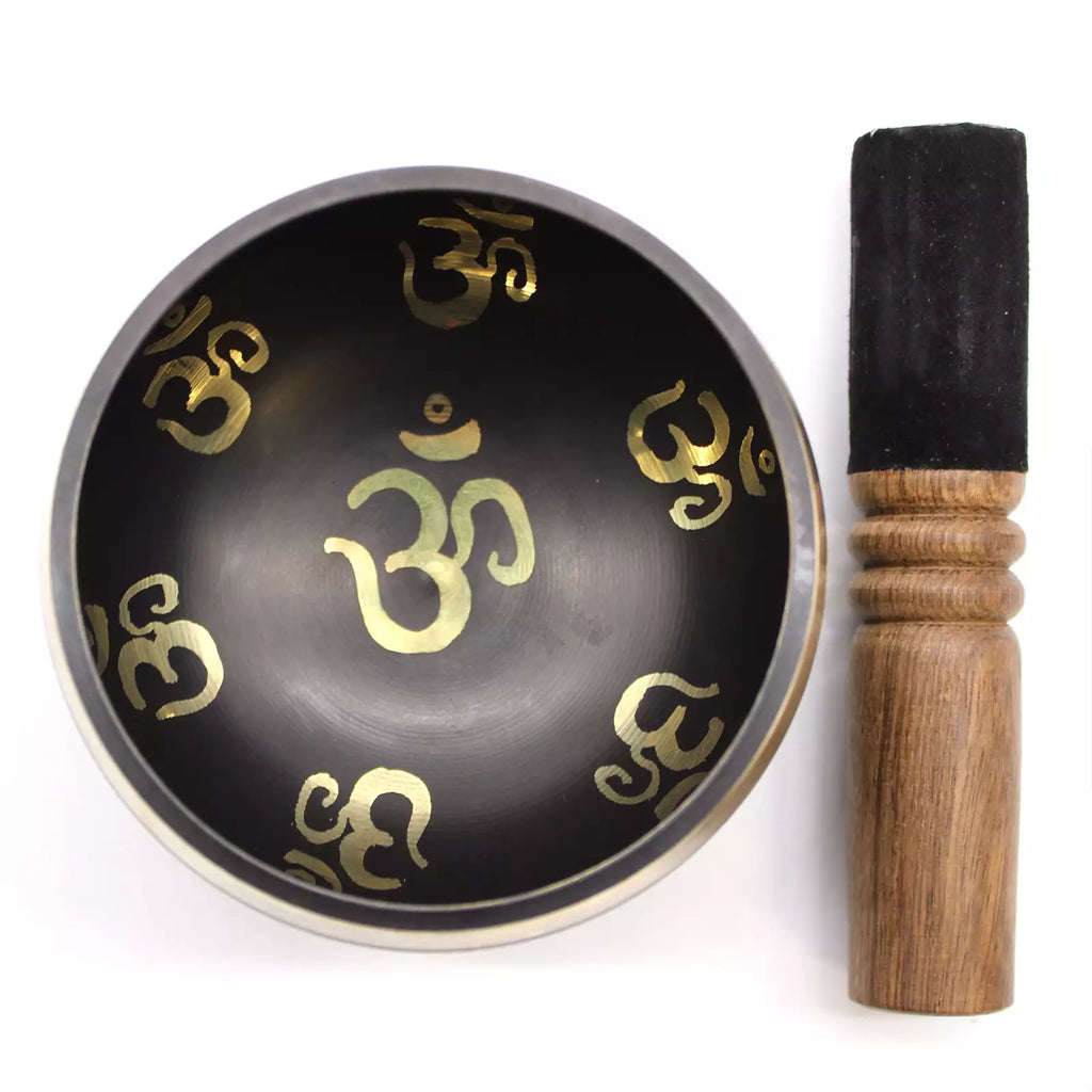 A black metal singing bowl decorated with gold Om symbols, shown next to its wooden mallet with a black felt tip on a white background.