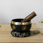 Black singing bowl with gold accents and a wooden striker on a wooden surface.