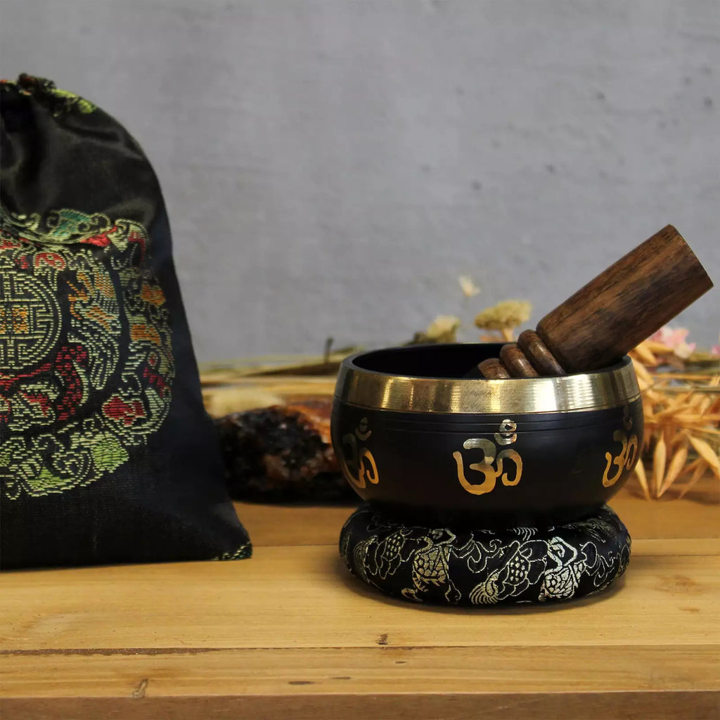 Black singing bowl with gold OM symbols on a wooden surface, accompanied by a wooden mallet and decorative bag.