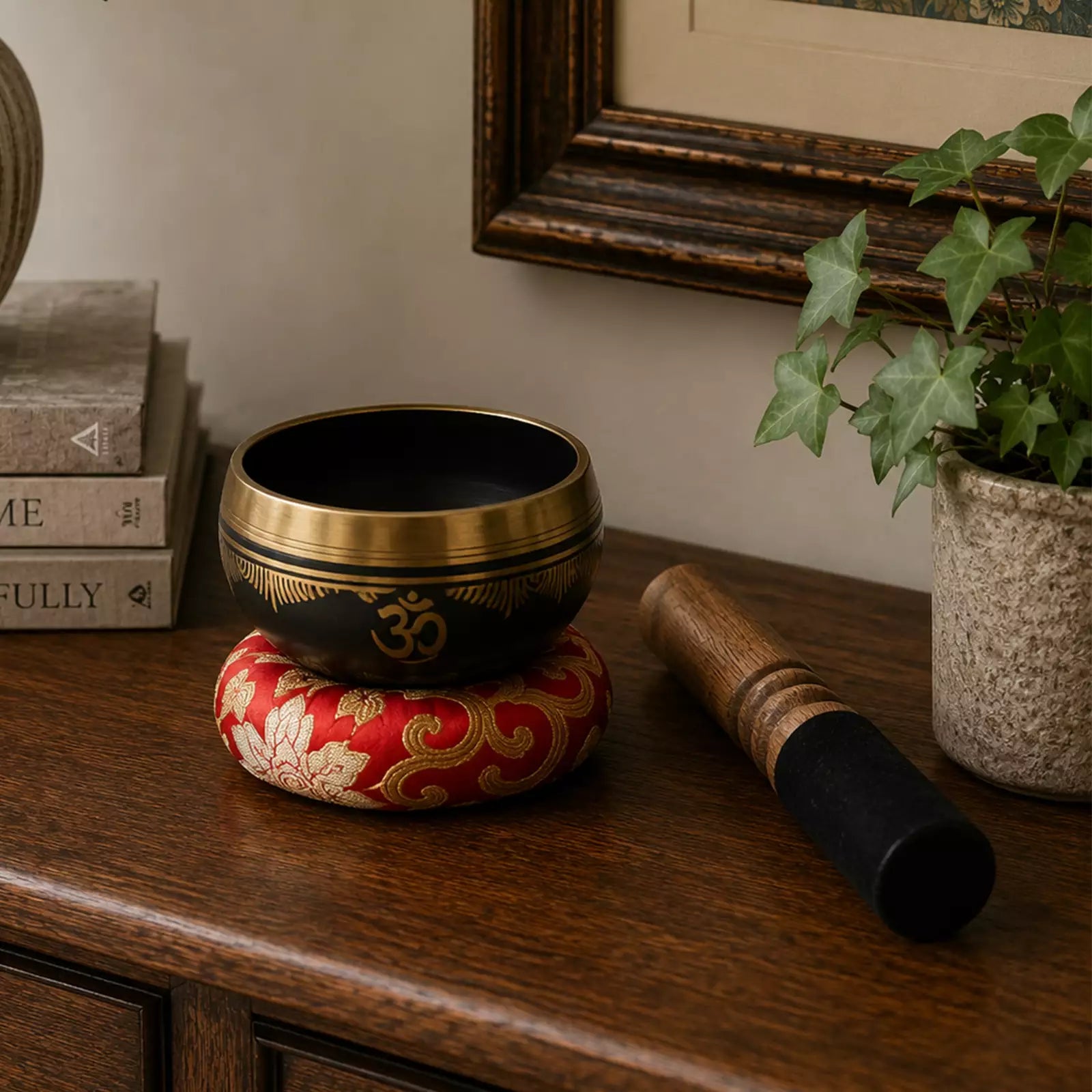 A black and gold Tibetan singing bowl with an Om symbol, resting on a red and gold patterned cushion, set on a wooden table next to a wooden mallet and a potted ivy plant.