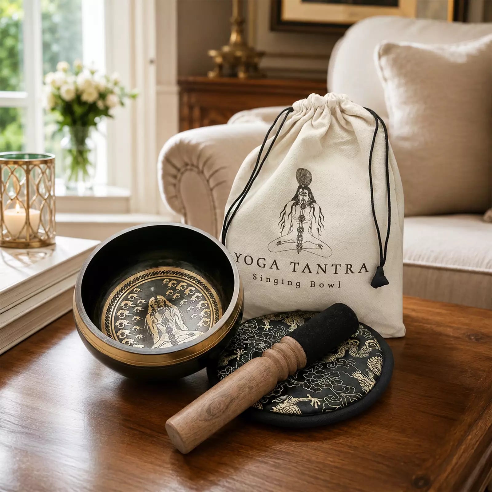 A singing bowl set displayed on a wooden table in a cozy living room setting. The set includes a black and gold Tibetan singing bowl, a wooden mallet, a black and gold carrying pouch printed with 'YOGA TANTRA Singing Bowl', and a black felt-covered striker.