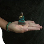A person wearing a dark sweater holds a Mini Hand Up Sitting Buddha statue with turquoise and gold coloring in their open palm, showcasing the small size and intricate details.
