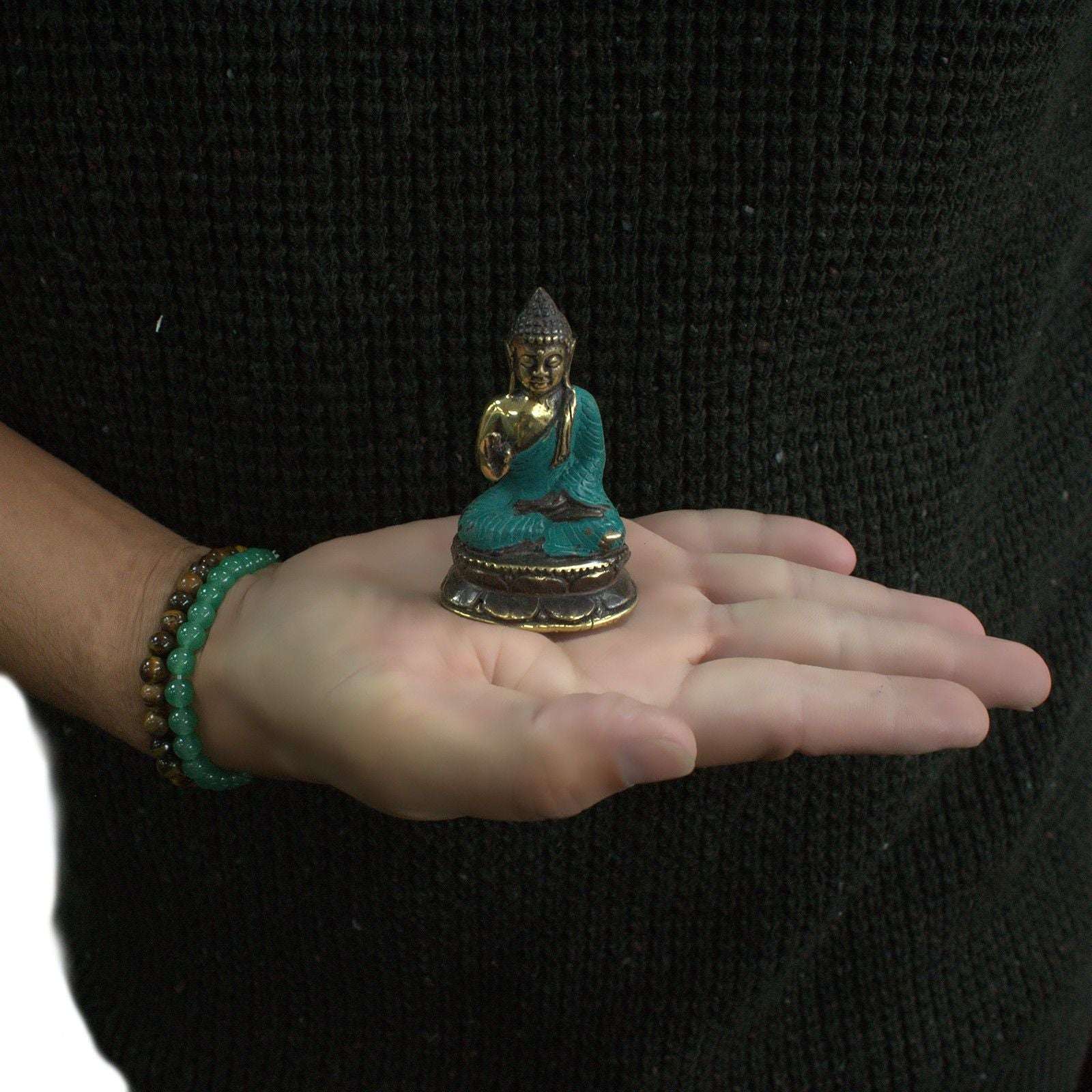 A person wearing a dark sweater holds a Mini Hand Up Sitting Buddha statue with turquoise and gold coloring in their open palm, showcasing the small size and intricate details.