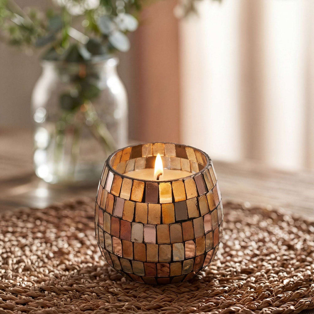 Small Glass Jar Candles - Amber Mosaic glowing with a warm flame, sitting on a woven surface with a blurred vase of greenery in the background.