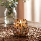 Small Glass Jar Candles - Amber Mosaic glowing with a warm flame, sitting on a woven surface with a blurred vase of greenery in the background.
