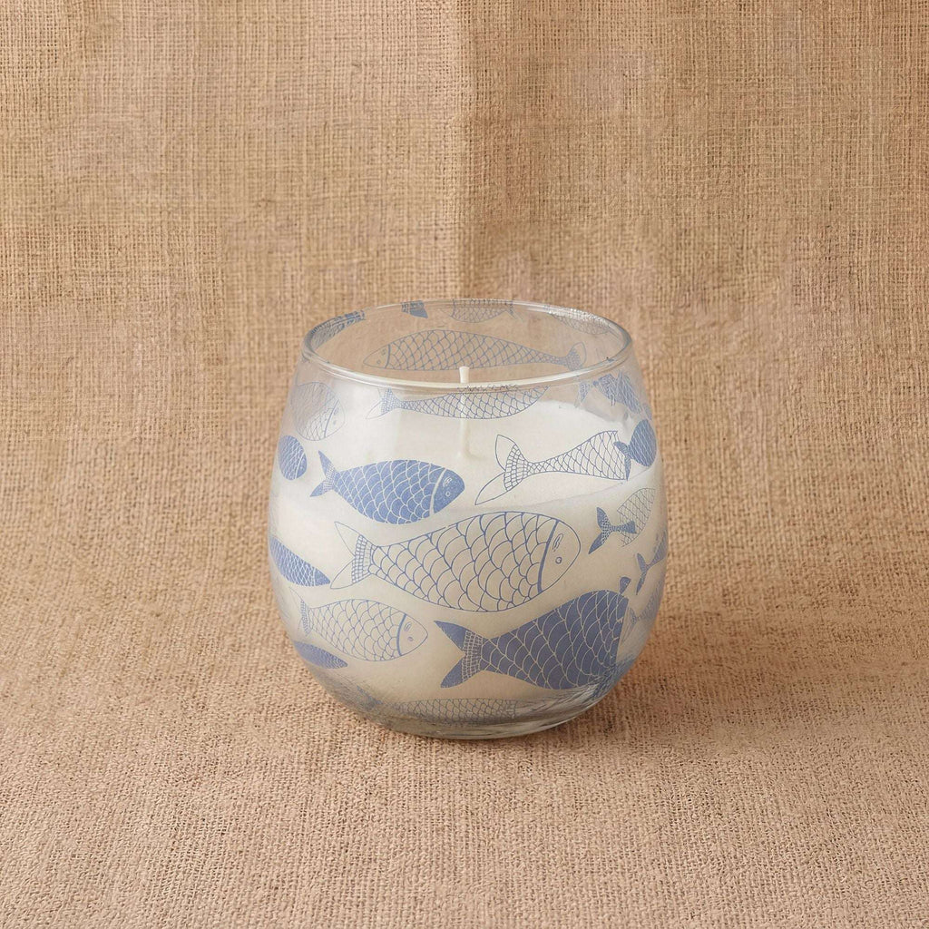 Small Glass Jar Candles - Seabreeze featuring a white candle in a clear glass jar decorated with blue fish illustrations, set on a textured beige fabric background.