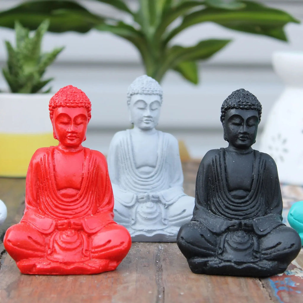 Three Matt Mini Buddha (Assorted Colours) statues in red, white, and black resin finish, displayed on a wooden surface with green plants in the background.