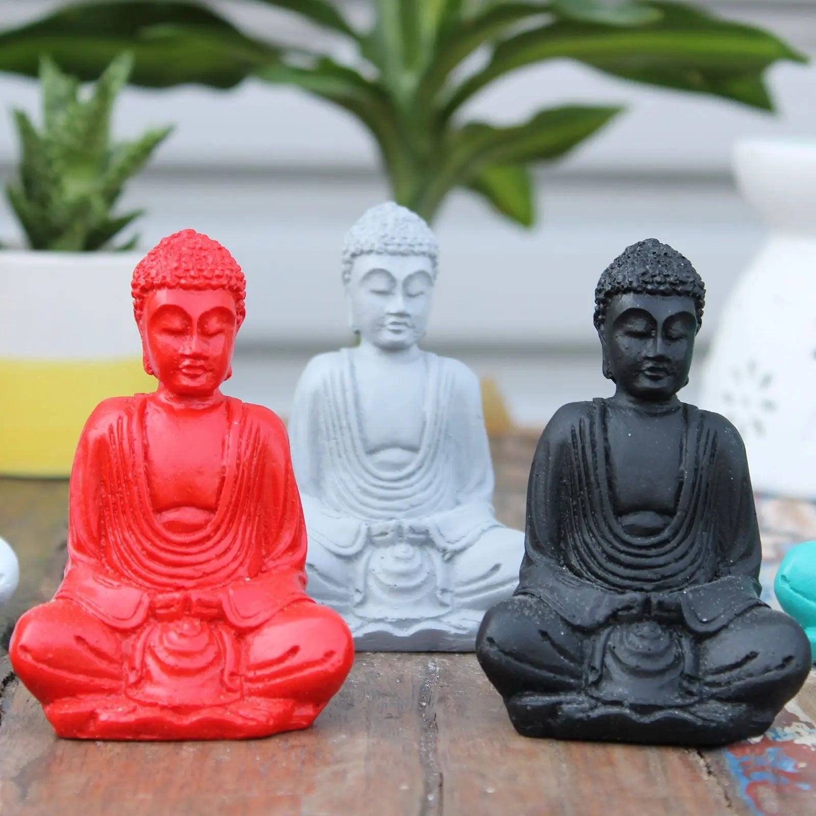 Three Matt Mini Buddha (Assorted Colours) statues in red, white, and black resin finish, displayed on a wooden surface with green plants in the background.