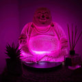 Laughing Buddha Lamp Dragonfruit - Round Stand glowing with a textured pink light, placed on a round base beside a small potted plant and reed diffuser, creating a calming ambiance.