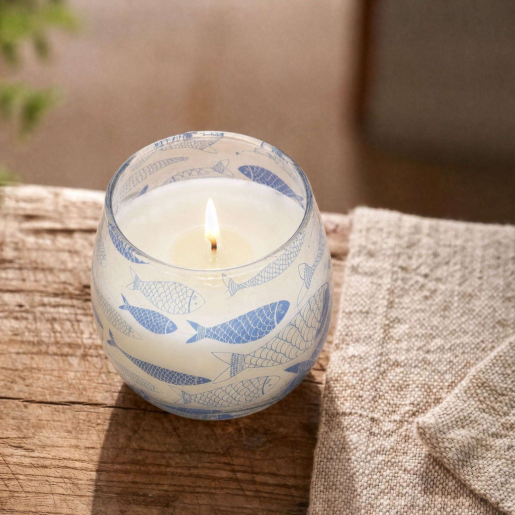 Small Glass Jar Candles - Seabreeze lit candle in a clear glass jar with blue fish designs, placed on a rustic wooden surface next to a beige textured fabric.