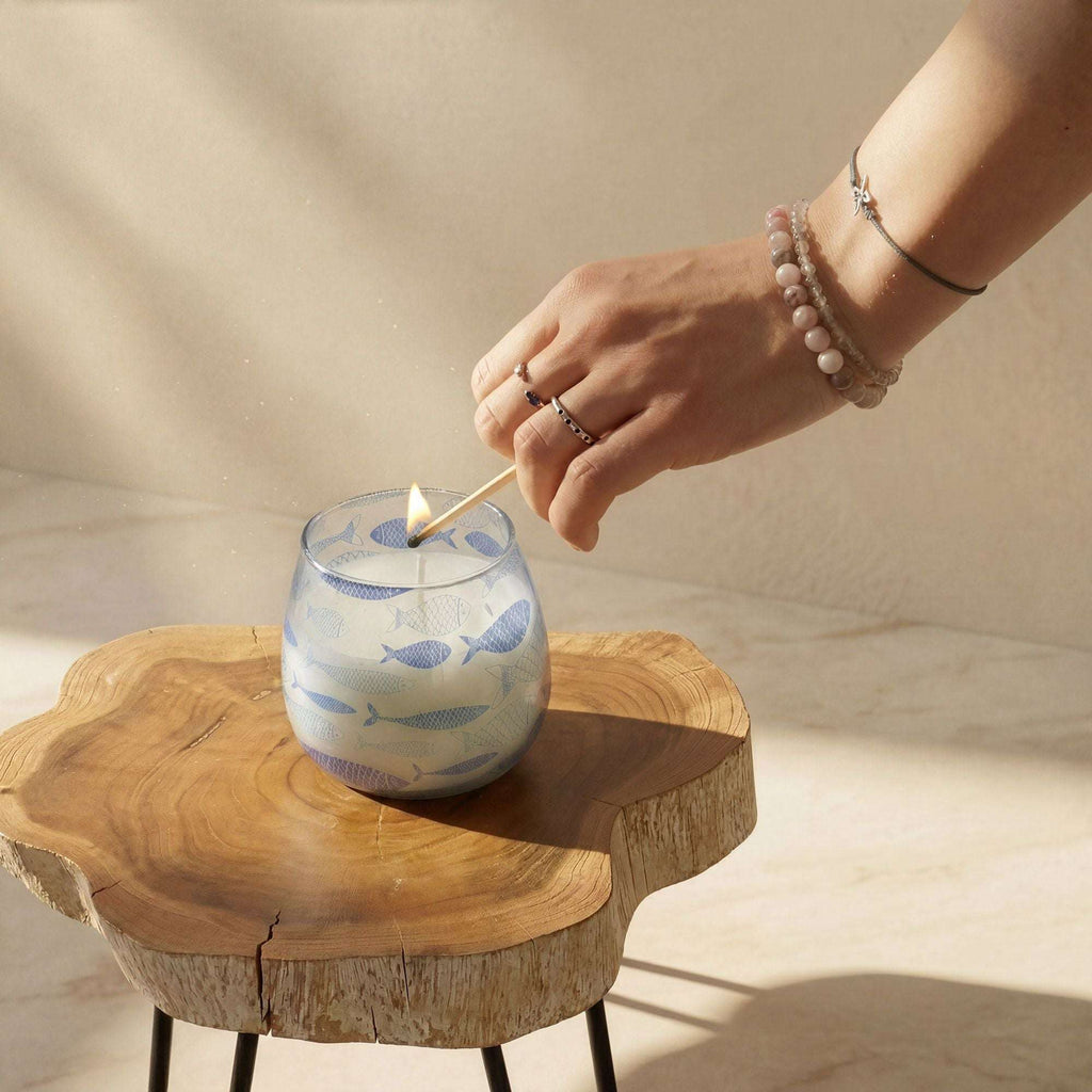Hand lighting a Small Glass Jar Candles - Seabreeze featuring a blue fish pattern, placed on a natural wooden surface in soft sunlight.