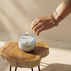 Hand lighting a Small Glass Jar Candles - Seabreeze featuring a blue fish pattern, placed on a natural wooden surface in soft sunlight.