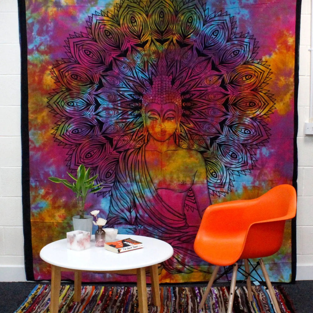 Vibrant Double Cotton Bedspread + Wall Hanging - Peaceful Buddha featuring a colorful tie-dye background and intricate black Buddha design, displayed behind a modern orange chair and white table.