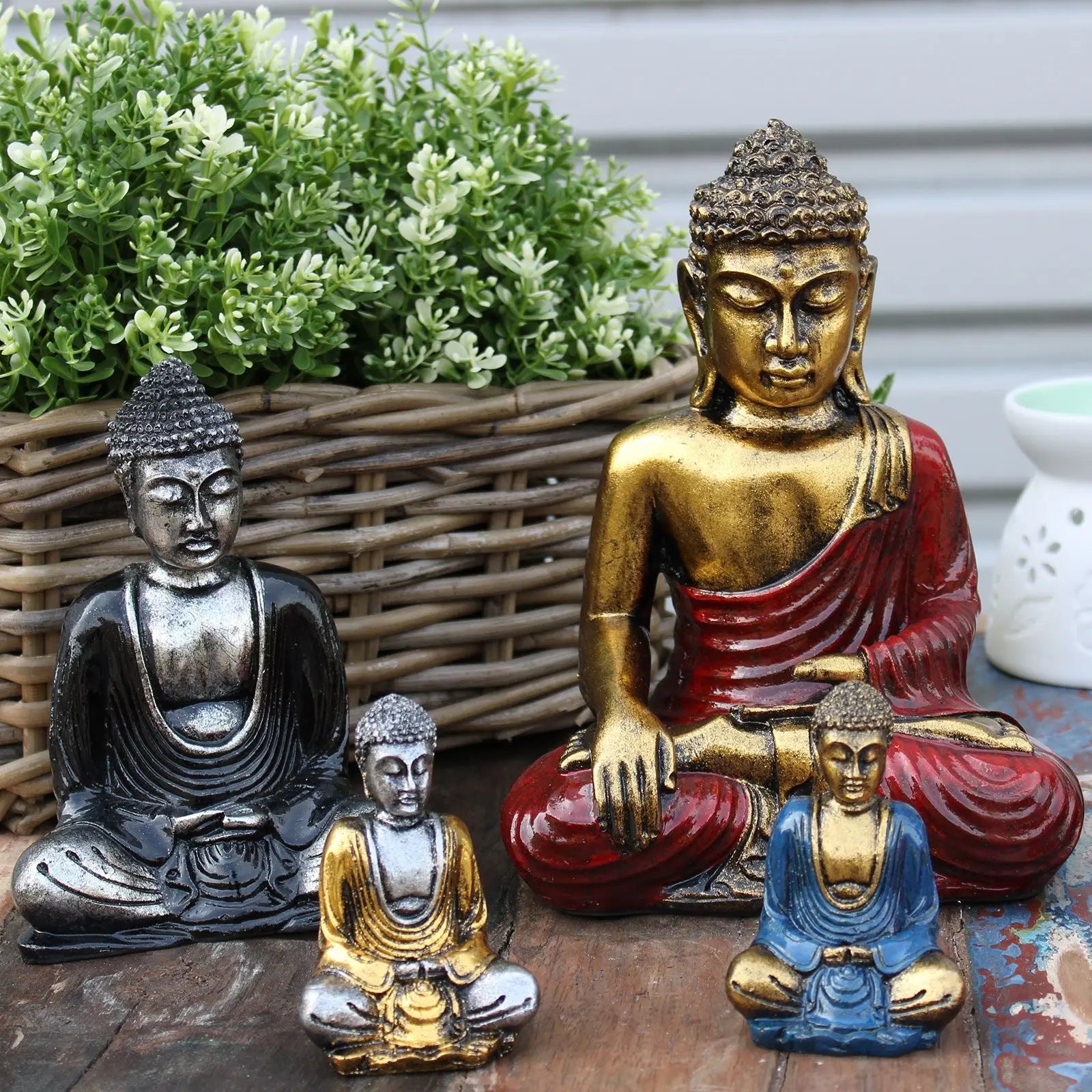 Red & Gold Buddha - Medium statue in a serene seated pose with gold and red finish, surrounded by smaller Buddha statues in metallic colors, placed on a rustic surface near a wicker basket with greenery.