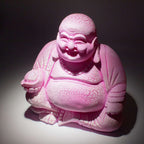 A pink cracked resin Laughing Buddha statue with a joyful expression, sitting on a round stand illuminated from above, featuring the product name Laughing Buddha Lamp Dragonfruit - Round Stand.