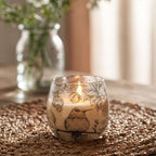 A lit Small Glass Jar Candles - Drawn by Flame with delicate black floral and bird illustrations on the glass, placed on a woven mat with a softly blurred background.