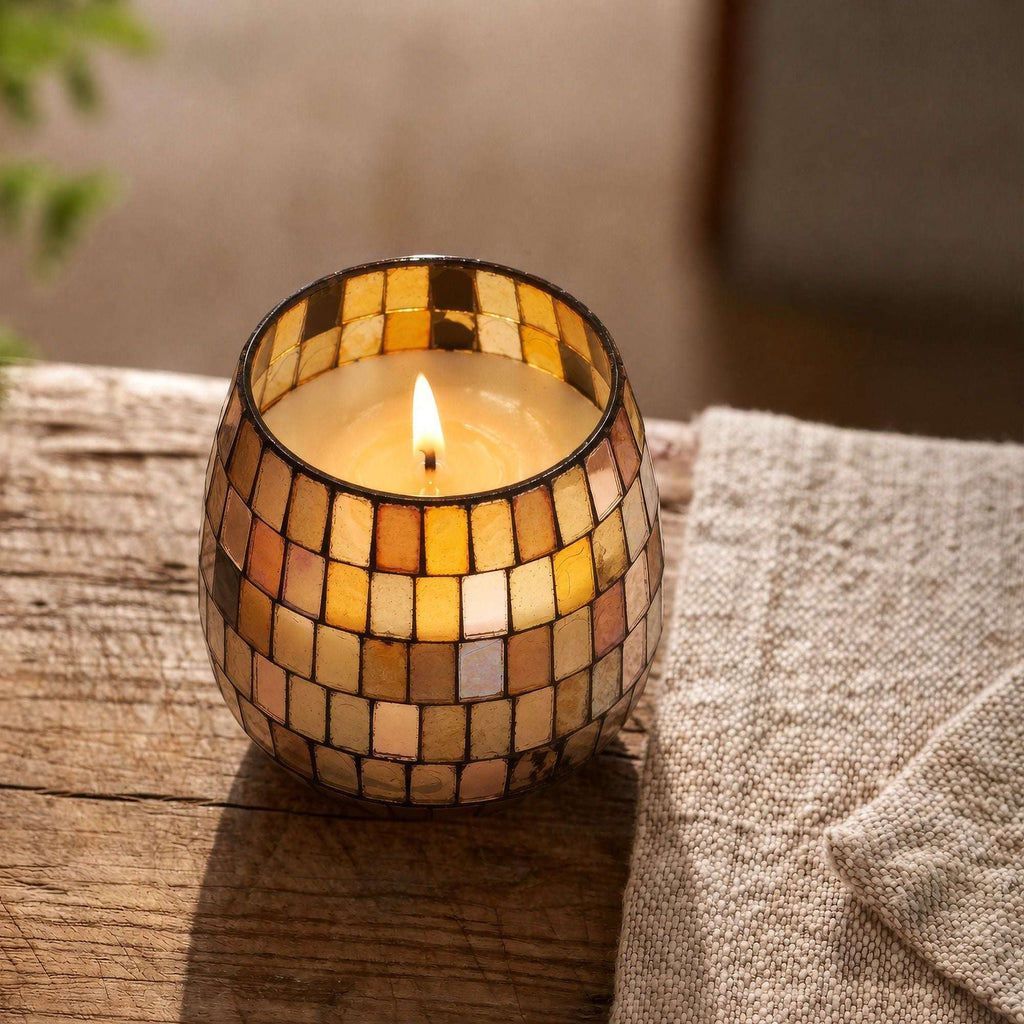 Small Glass Jar Candles - Amber Mosaic lit candle in a decorative amber mosaic glass jar placed on a rustic wooden surface next to a textured beige fabric.