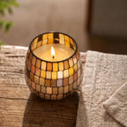 Small Glass Jar Candles - Amber Mosaic lit candle in a decorative amber mosaic glass jar placed on a rustic wooden surface next to a textured beige fabric.