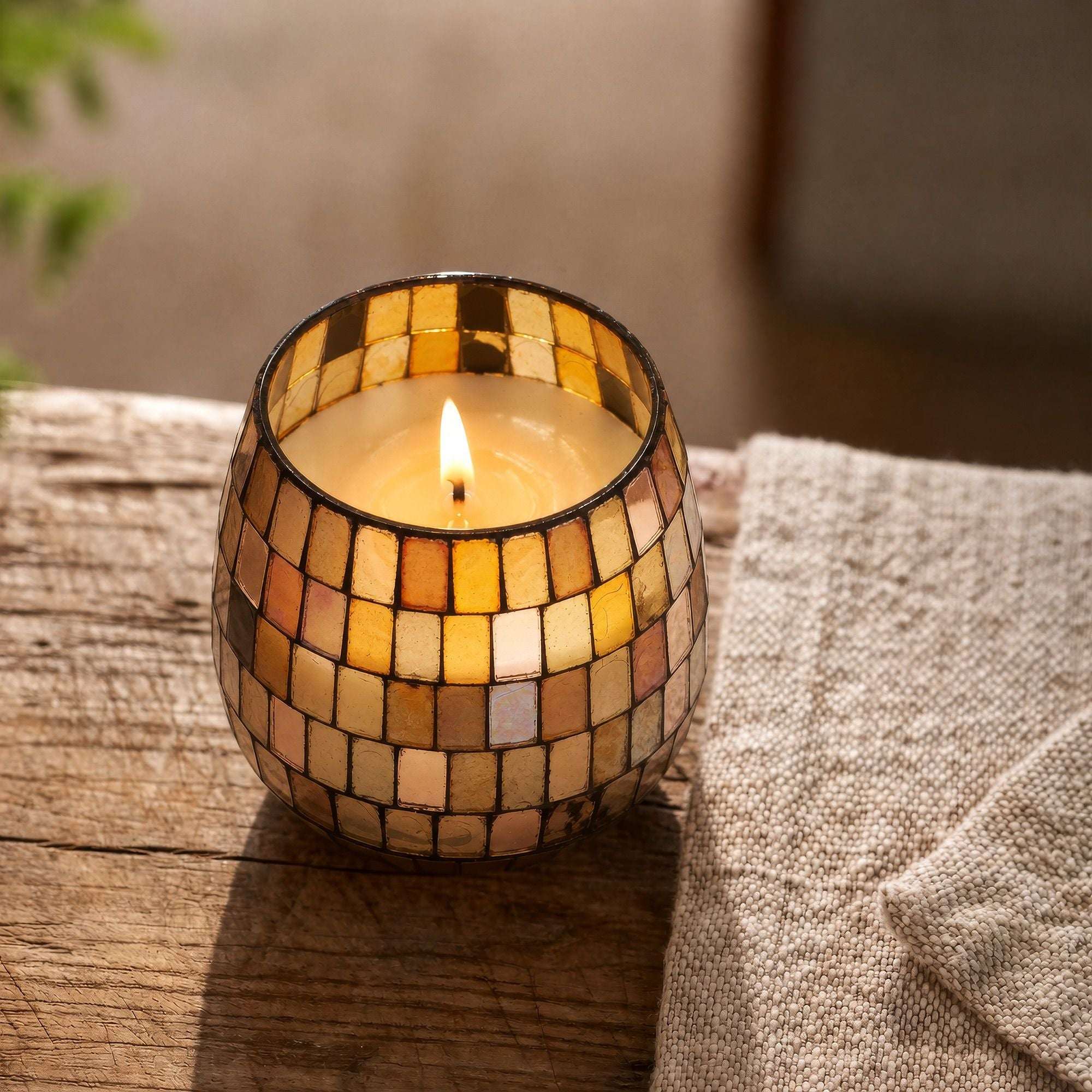 Small Glass Jar Candles - Amber Mosaic lit candle in a decorative amber mosaic glass jar placed on a rustic wooden surface next to a textured beige fabric.