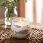 Medium Glass Jar Candles - Seabreeze with a lit wick inside a glass container decorated with blue fish patterns, placed on a woven surface in a softly lit room.